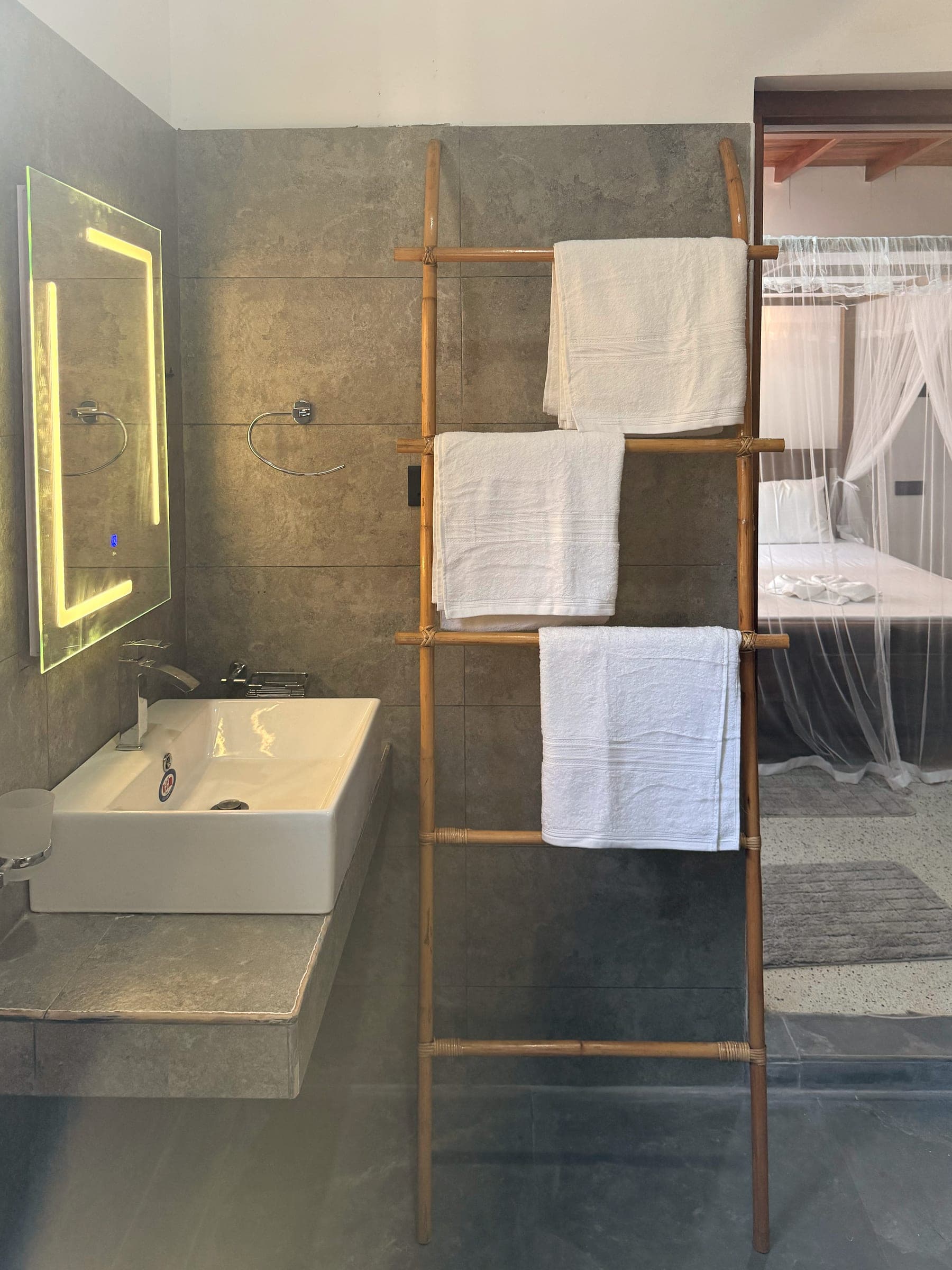 Bathroom with grey stone walls, a backlit mirror and a bamboo towel ladder, the bedroom beyond